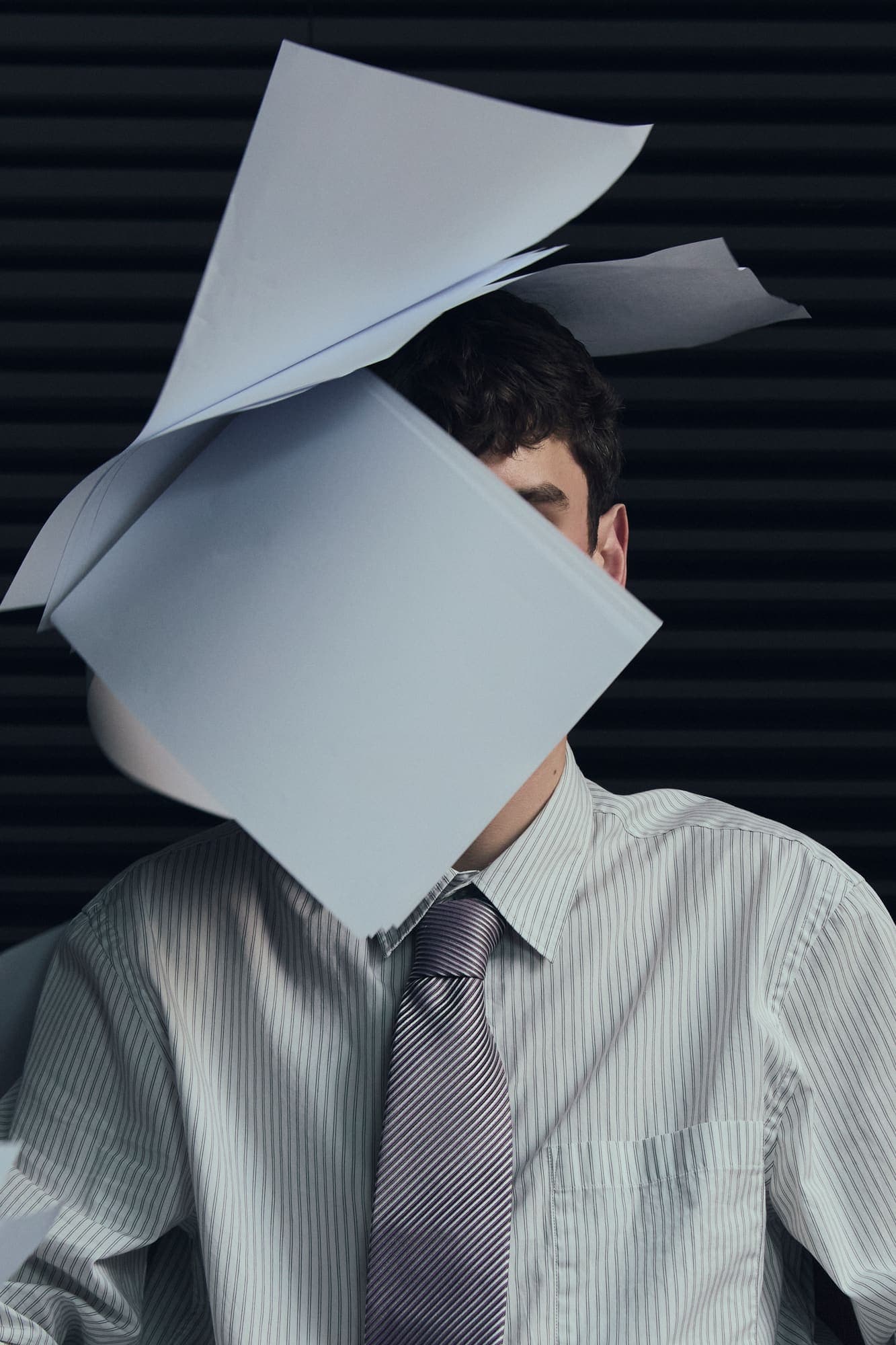 Sheets of paper piled over a seated man's head, hiding his face entirely