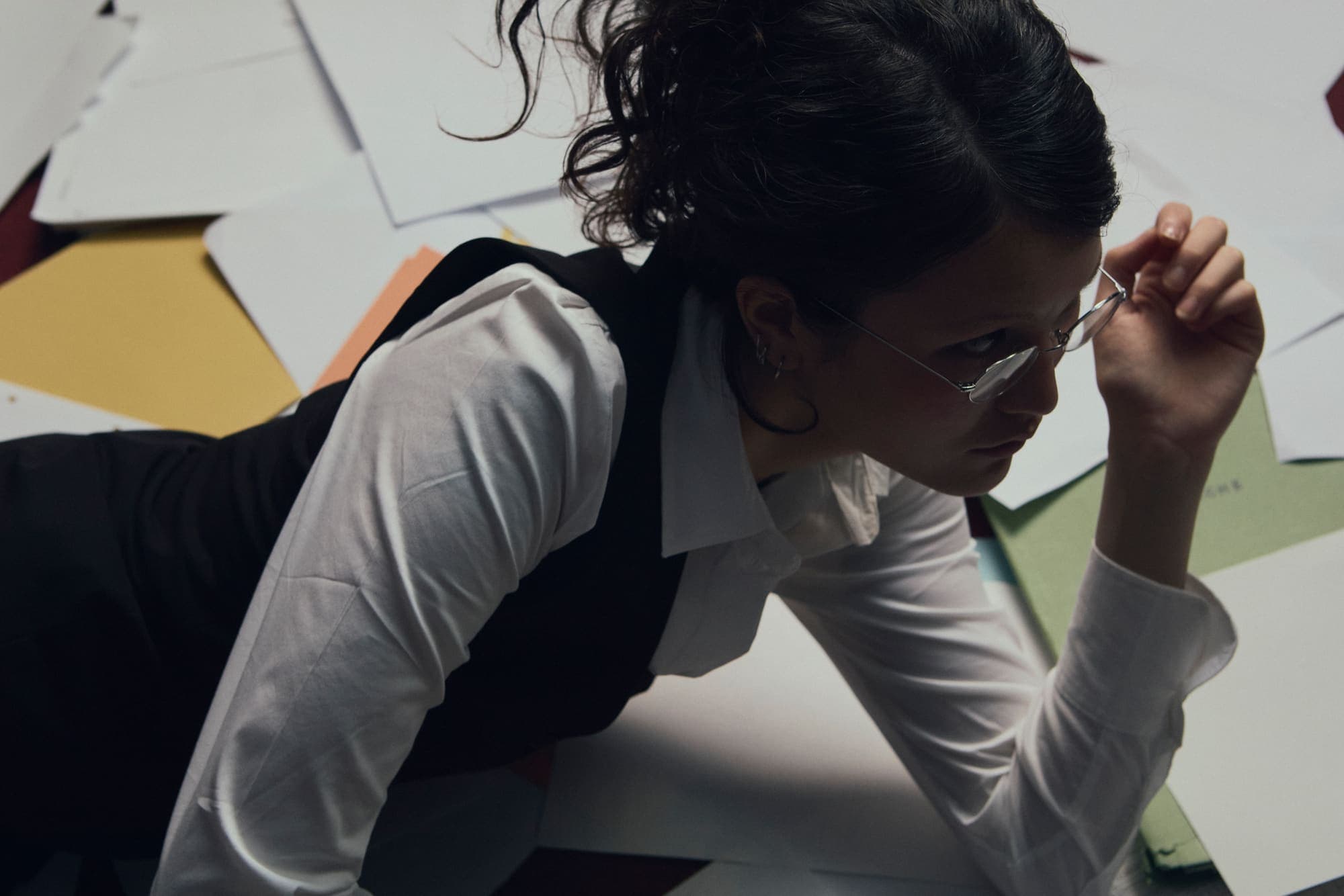 A woman adjusting her glasses while crawling across a floor of scattered papers