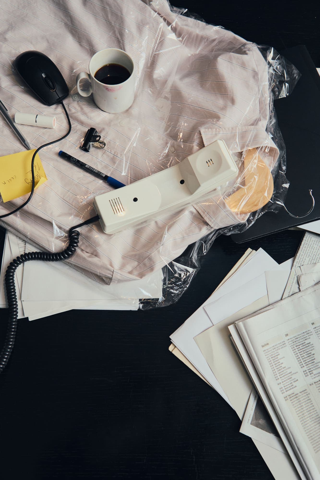 Overhead view of a cluttered desk with a coffee cup, phone handset, mouse and piles of paperwork