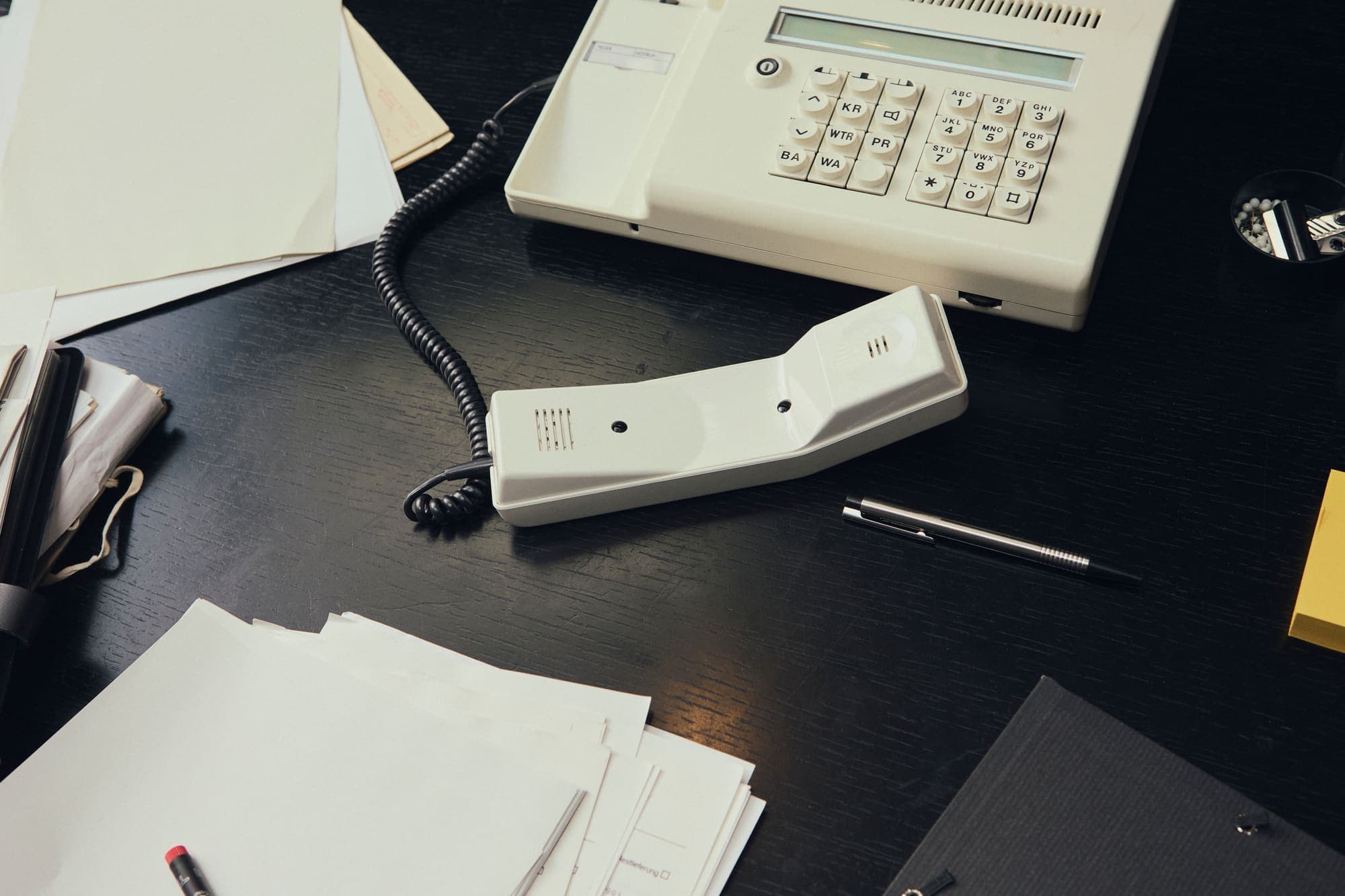 Overhead view of a desk phone off the hook among scattered papers and a pen on a dark desk