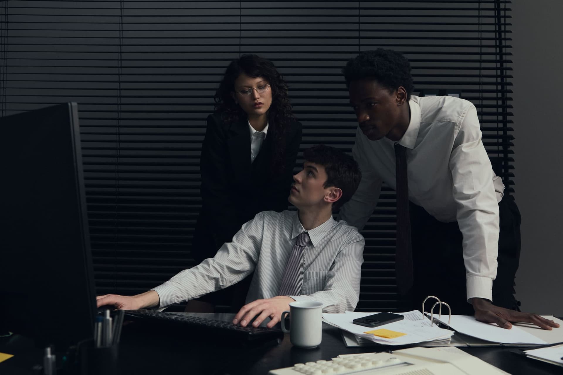 Three colleagues gathered around a monitor in a dark office, one seated and typing