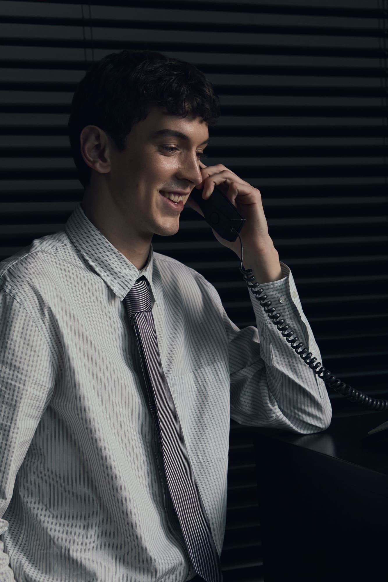 A smiling man taking a phone call at his desk in front of venetian blinds