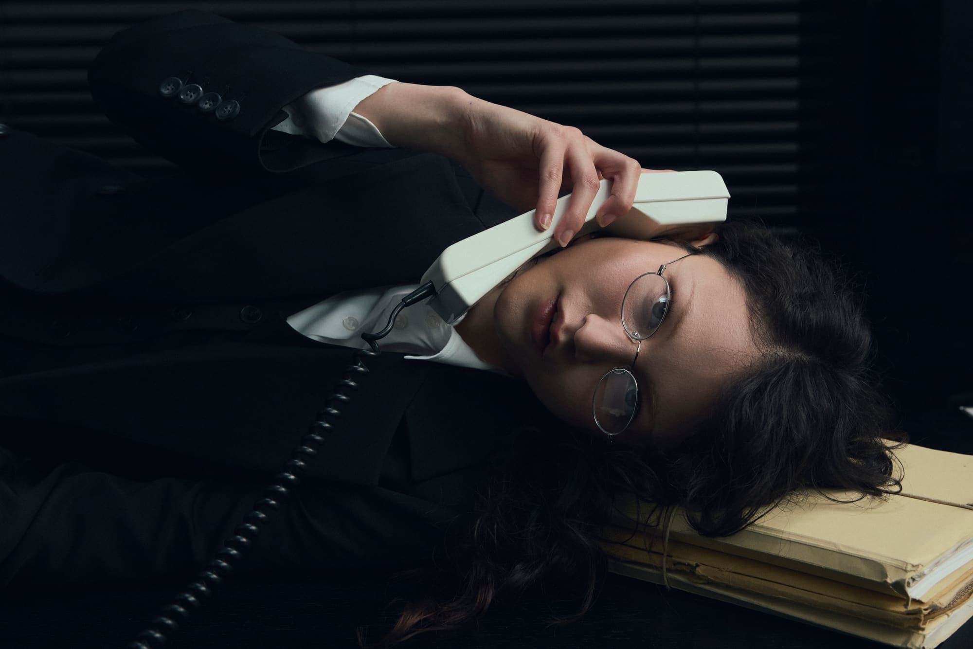 A woman lying back on stacked folders pressing a phone handset to her forehead