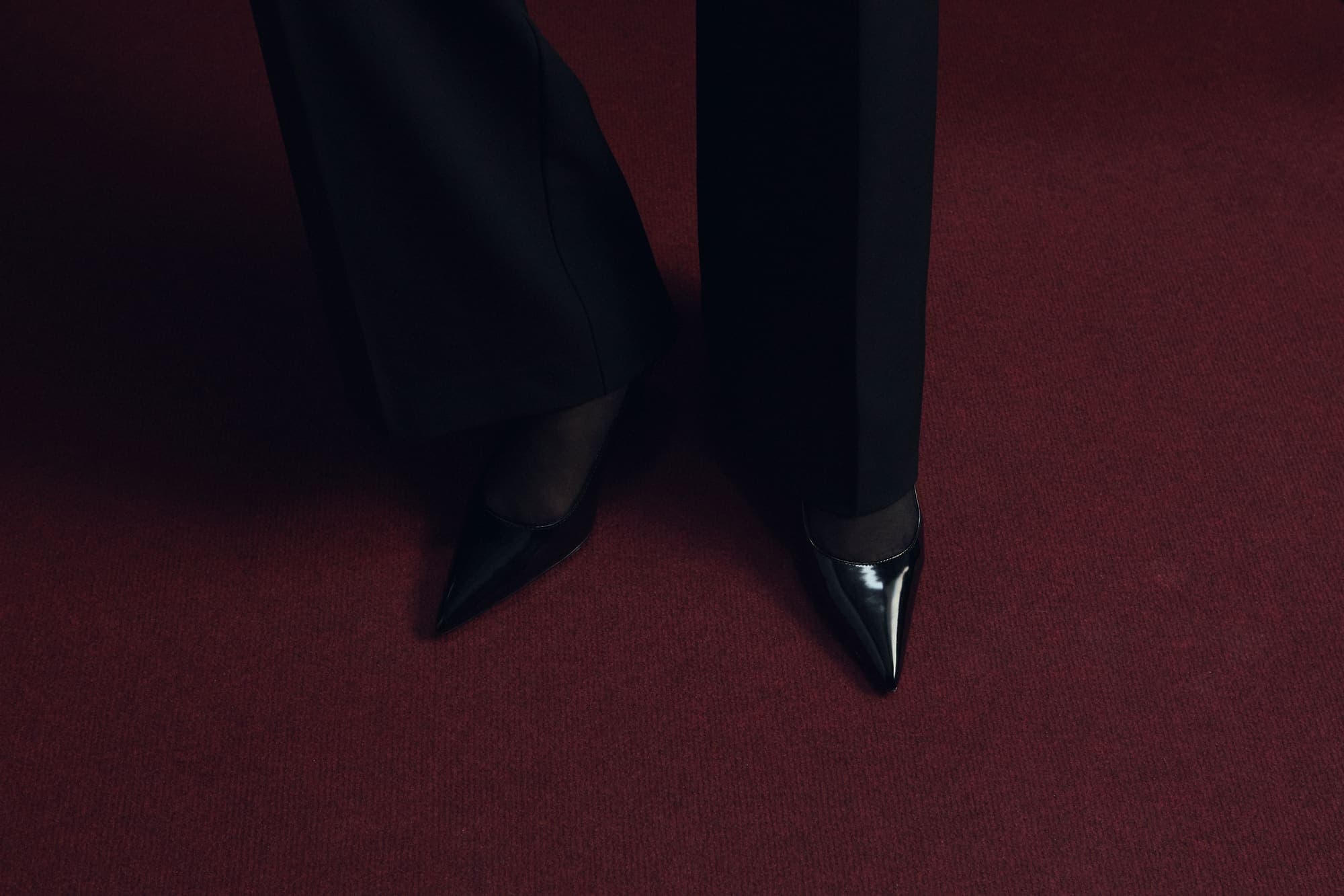 Pointed black heels and dark trousers standing on an oxblood-red carpet