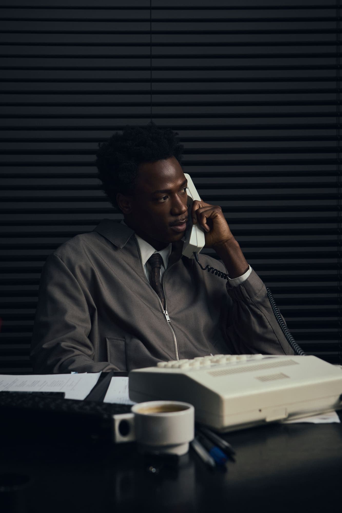 A man on a late-night phone call at a desk with a coffee cup and a beige desk phone