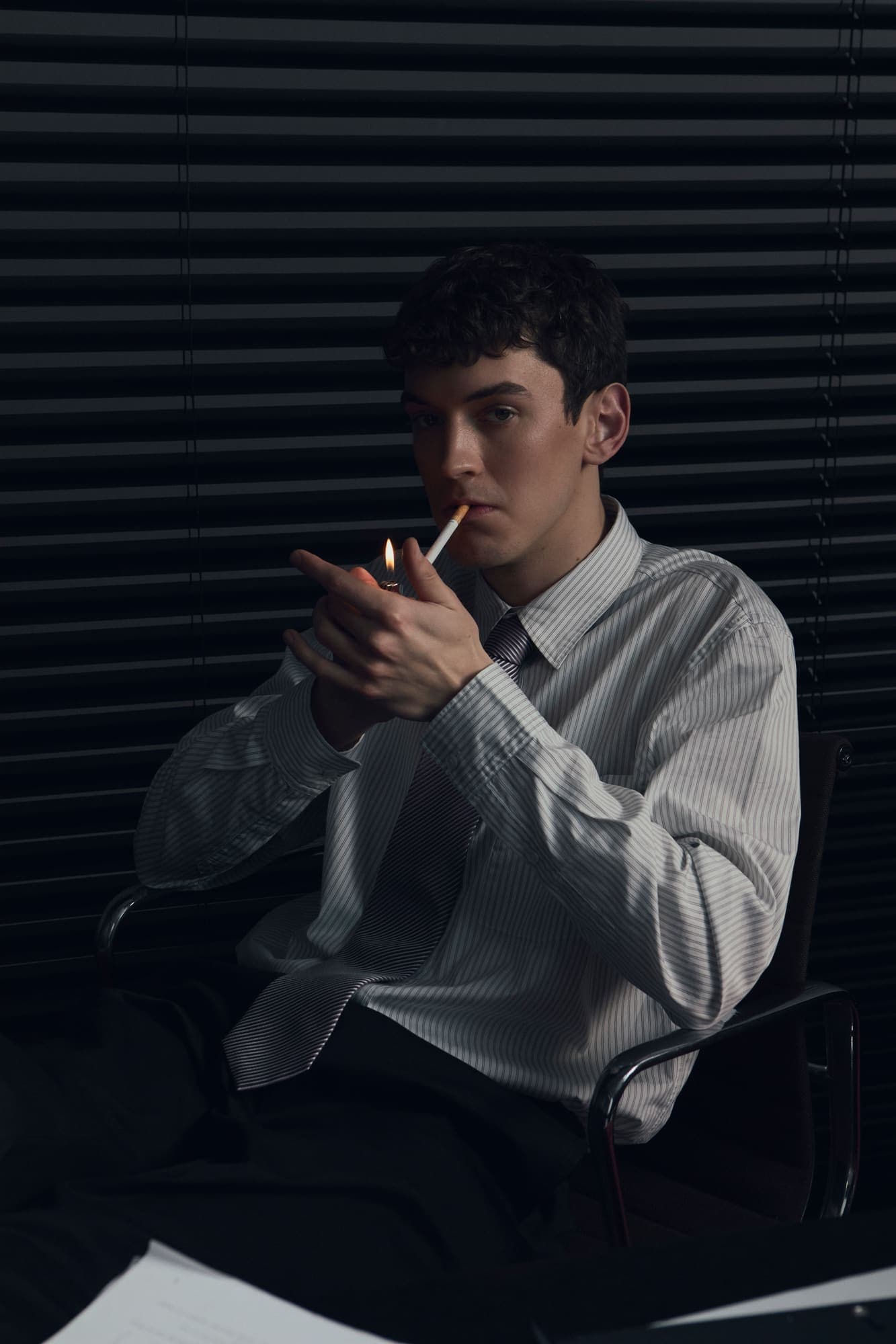 A man seated in an office chair lighting a cigarette in a dark room