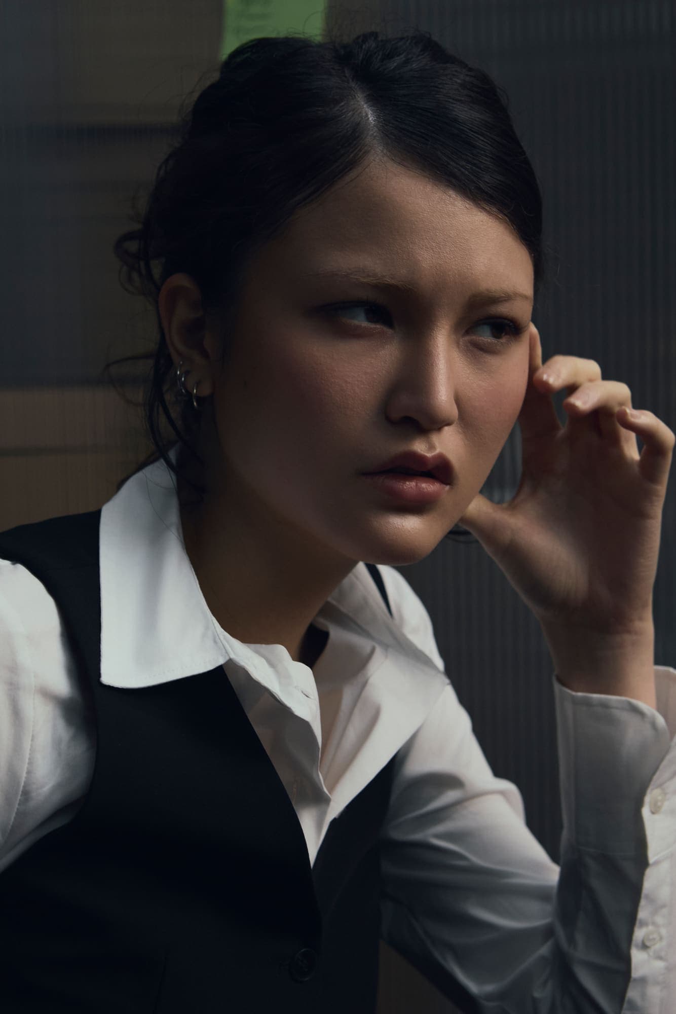 A woman in a dark vest touching her ear and glancing sideways