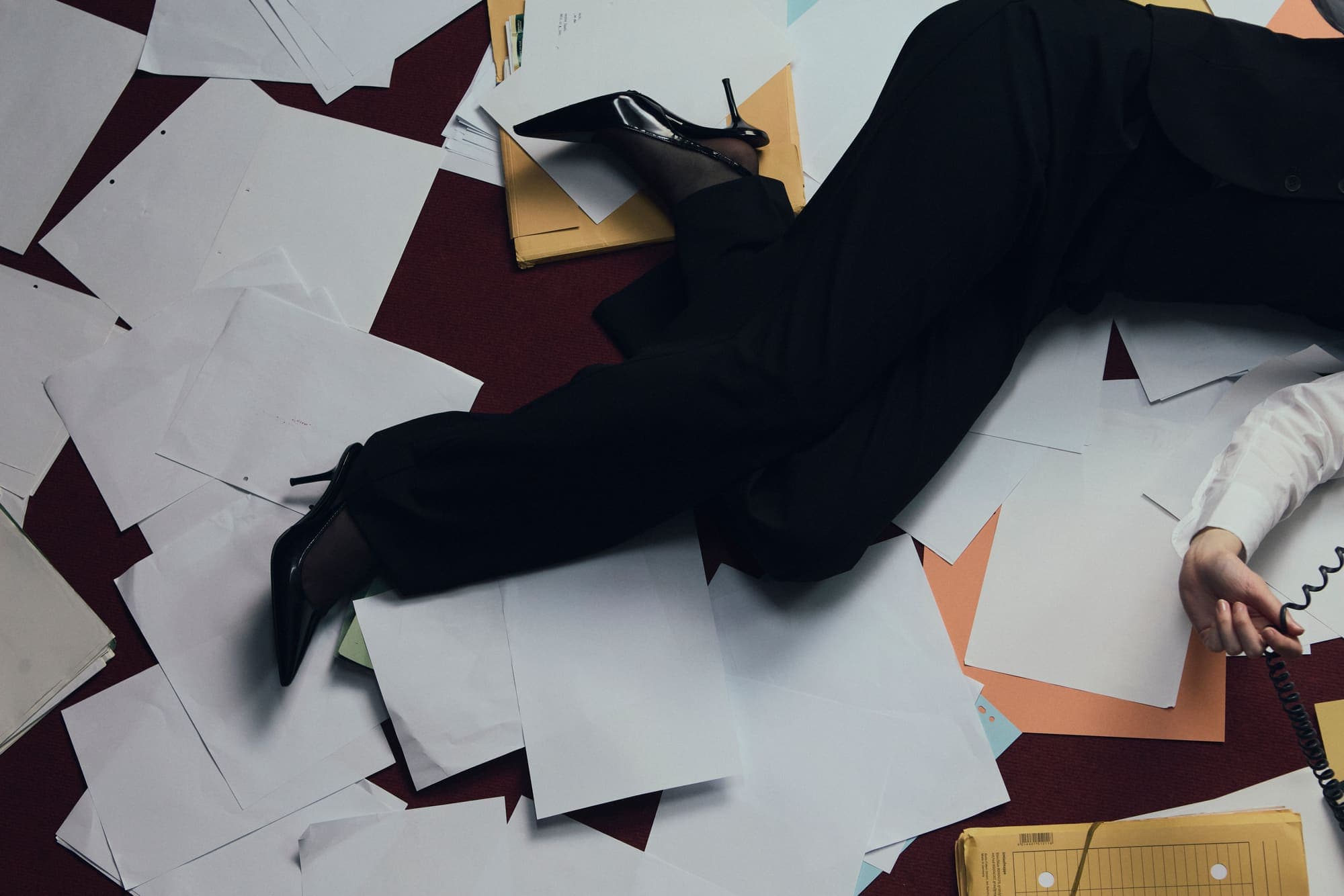 A person lying across a paper-strewn floor reaching for a phone cord