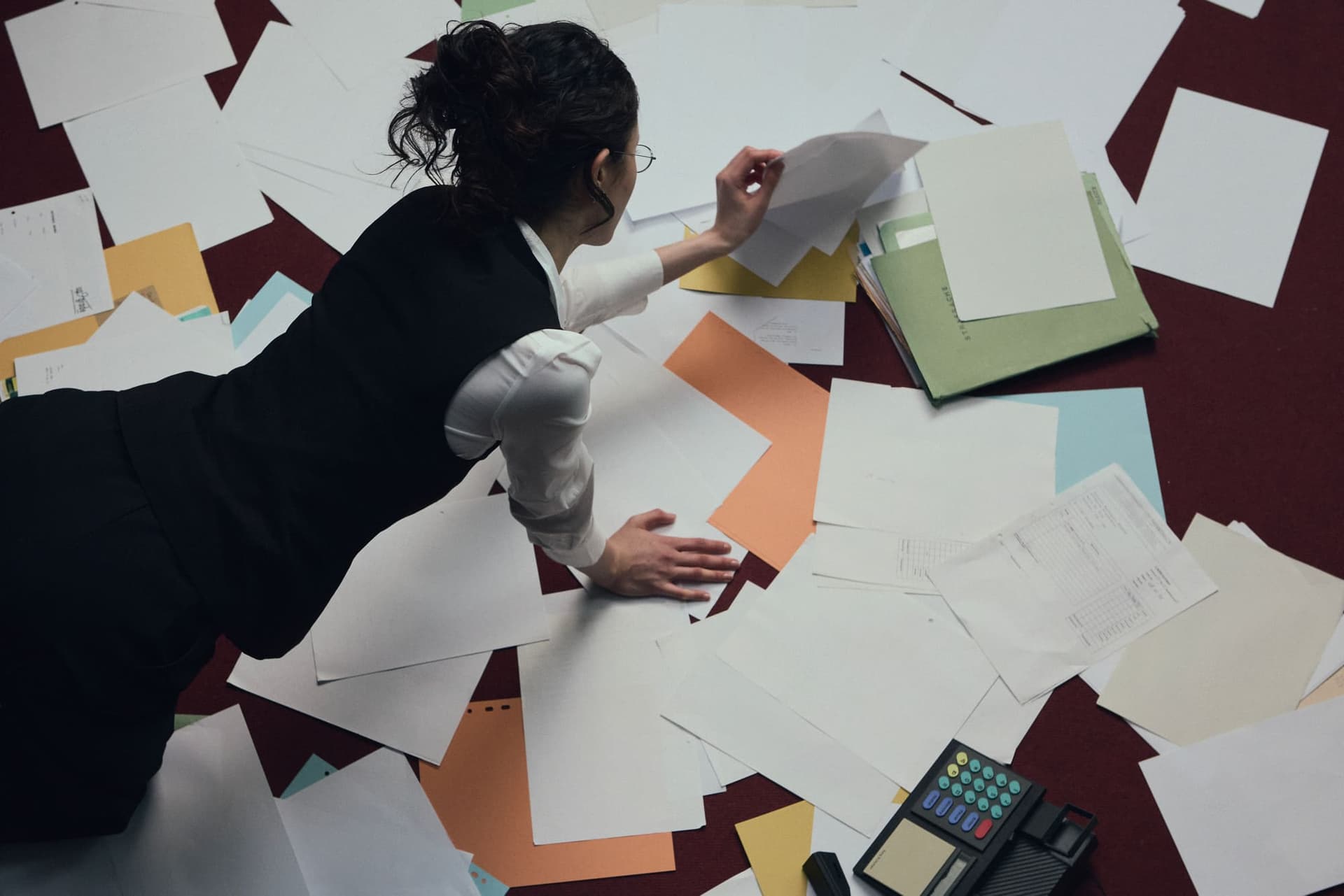 A woman crawling across a floor completely covered in scattered papers, reaching for a page