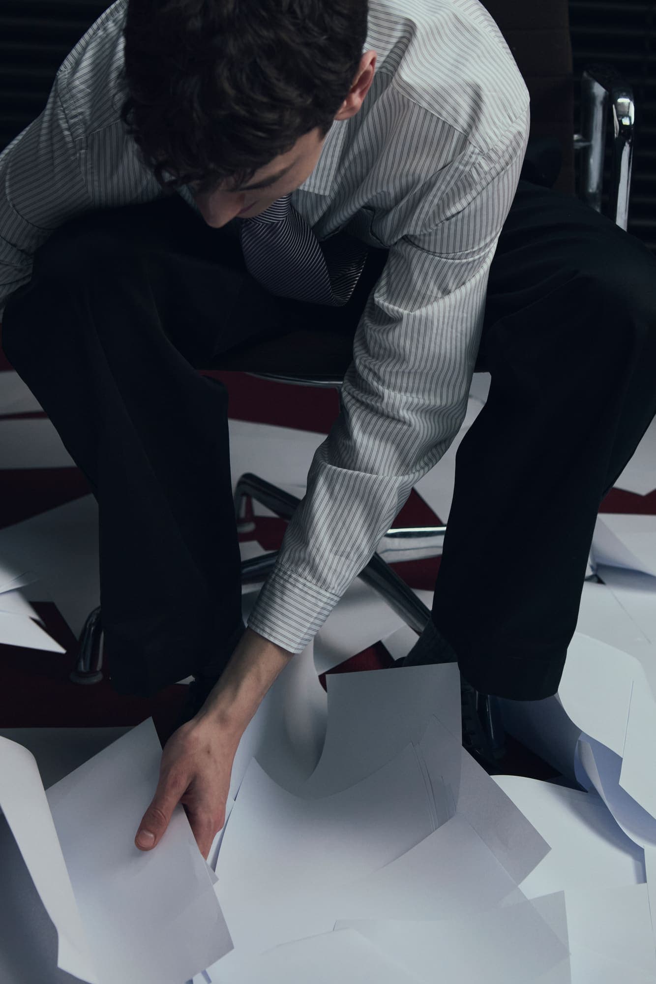 A man leaning down from his chair to gather papers scattered across red carpet