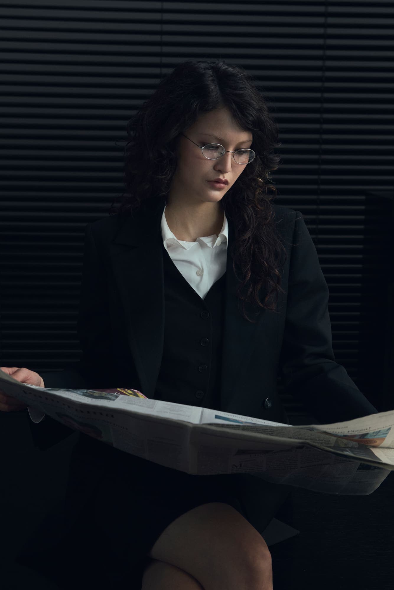 A woman in a dark suit reading a newspaper in front of venetian blinds