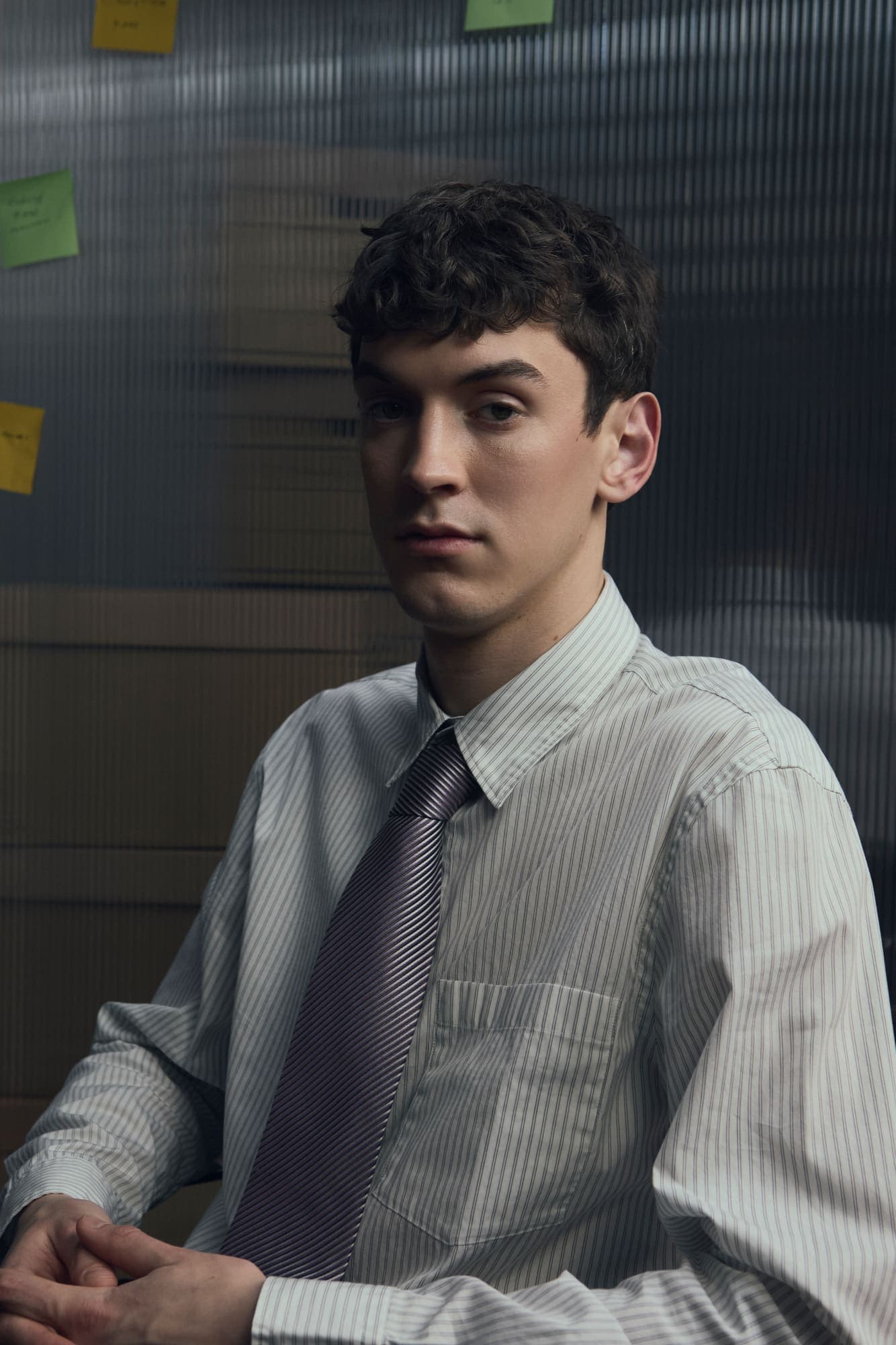 A young man in shirt and tie seated calmly in front of stacked boxes and reeded glass