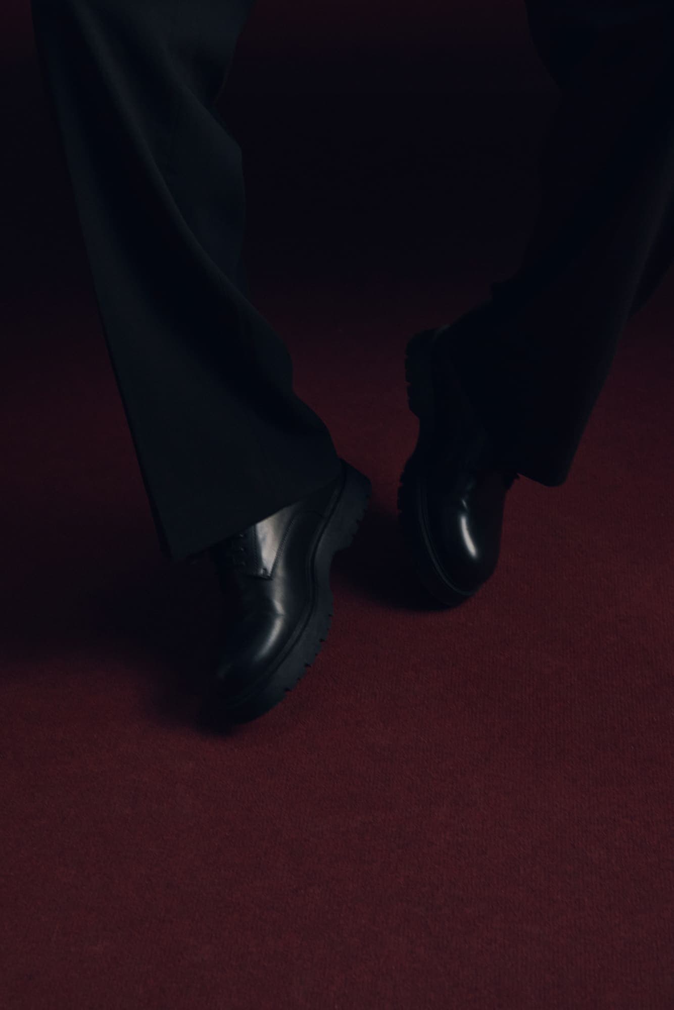 Polished black shoes and dark trousers mid-step on an oxblood-red carpet