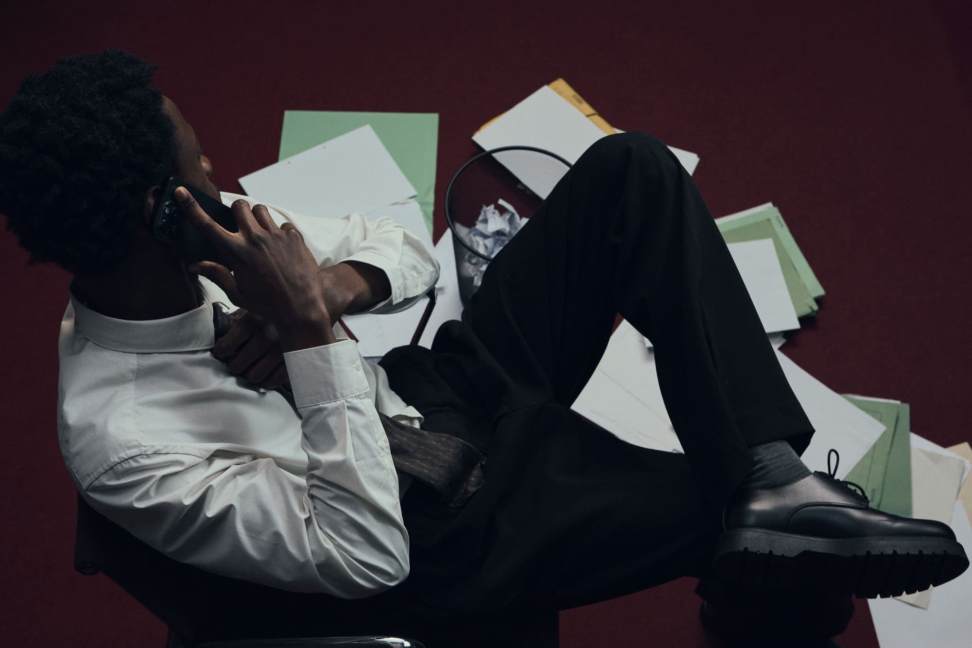 A man sprawled across a chair taking a phone call above papers scattered on red carpet