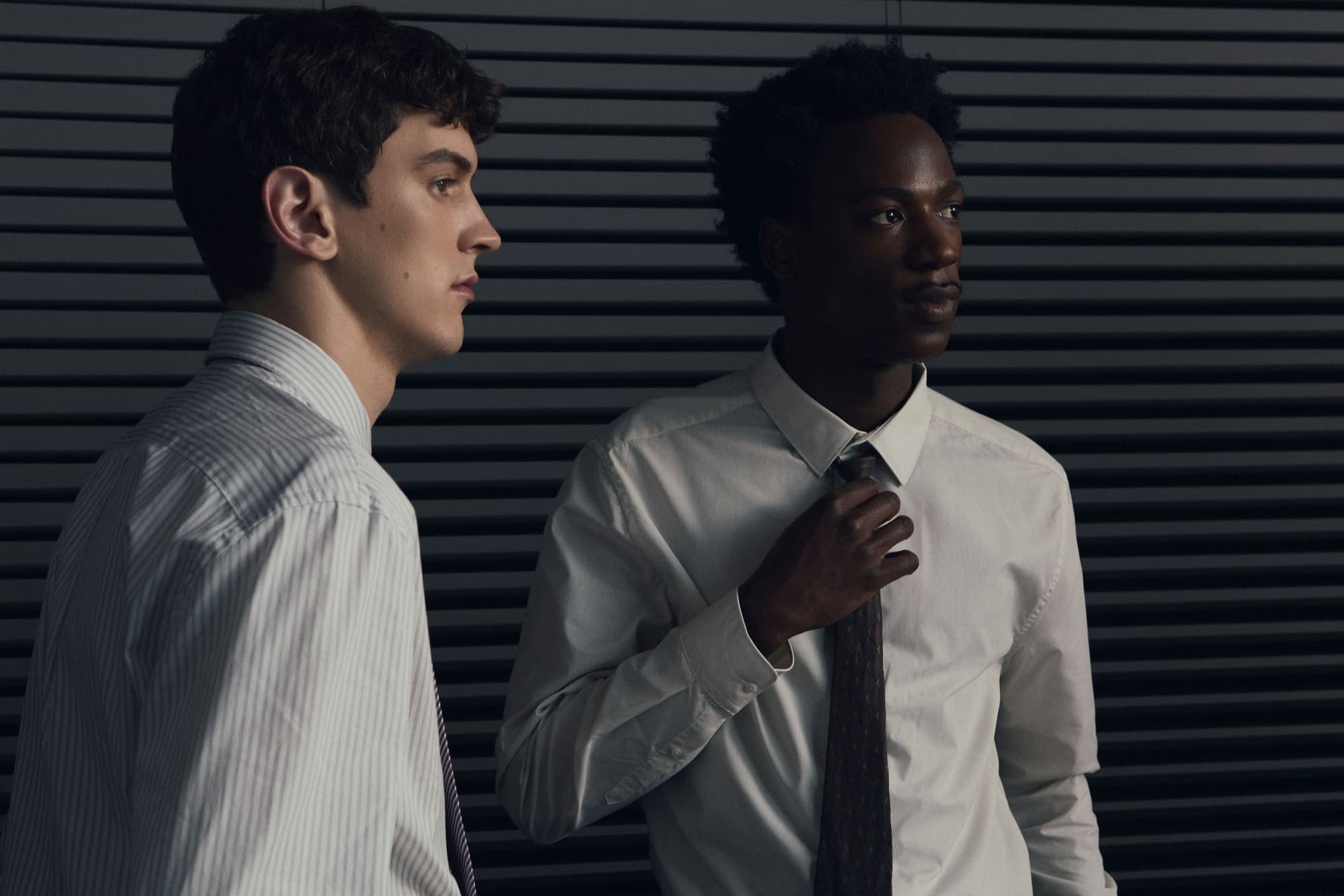 Two colleagues in shirts and ties standing before venetian blinds, one adjusting his tie