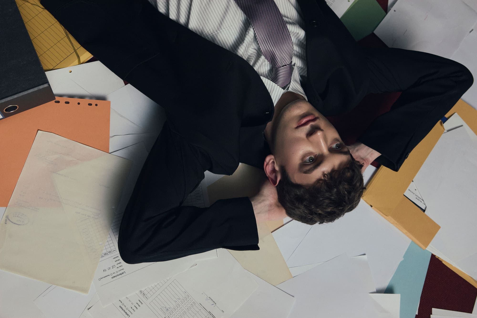 A man in a suit lying upside down across a paper-scattered floor