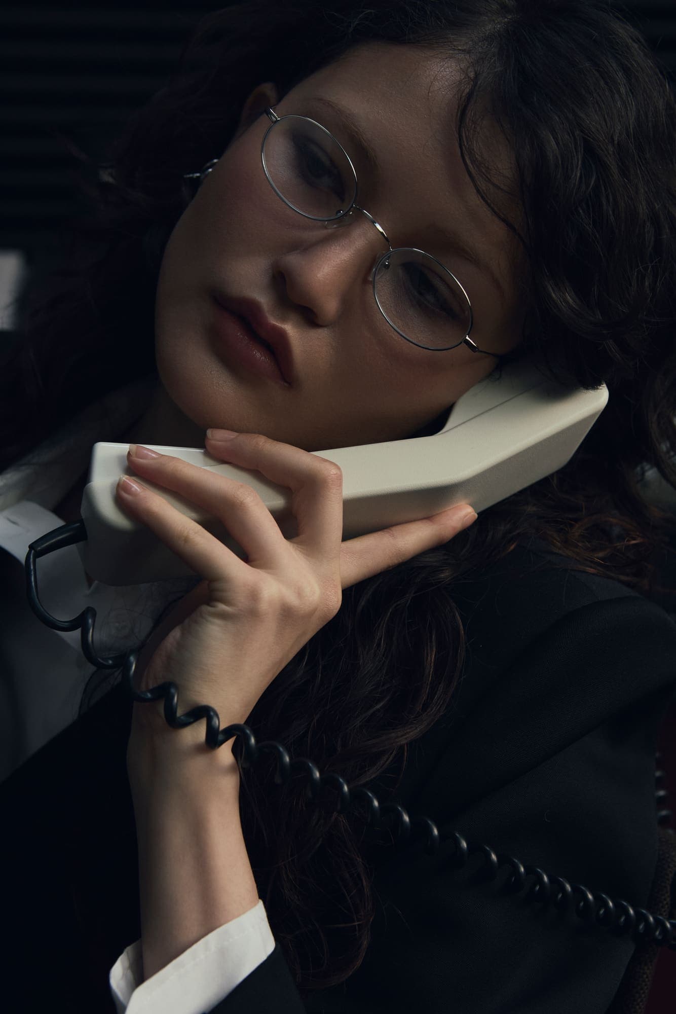 A woman in round glasses pressing a beige handset to her ear with the cord wrapped around her wrist