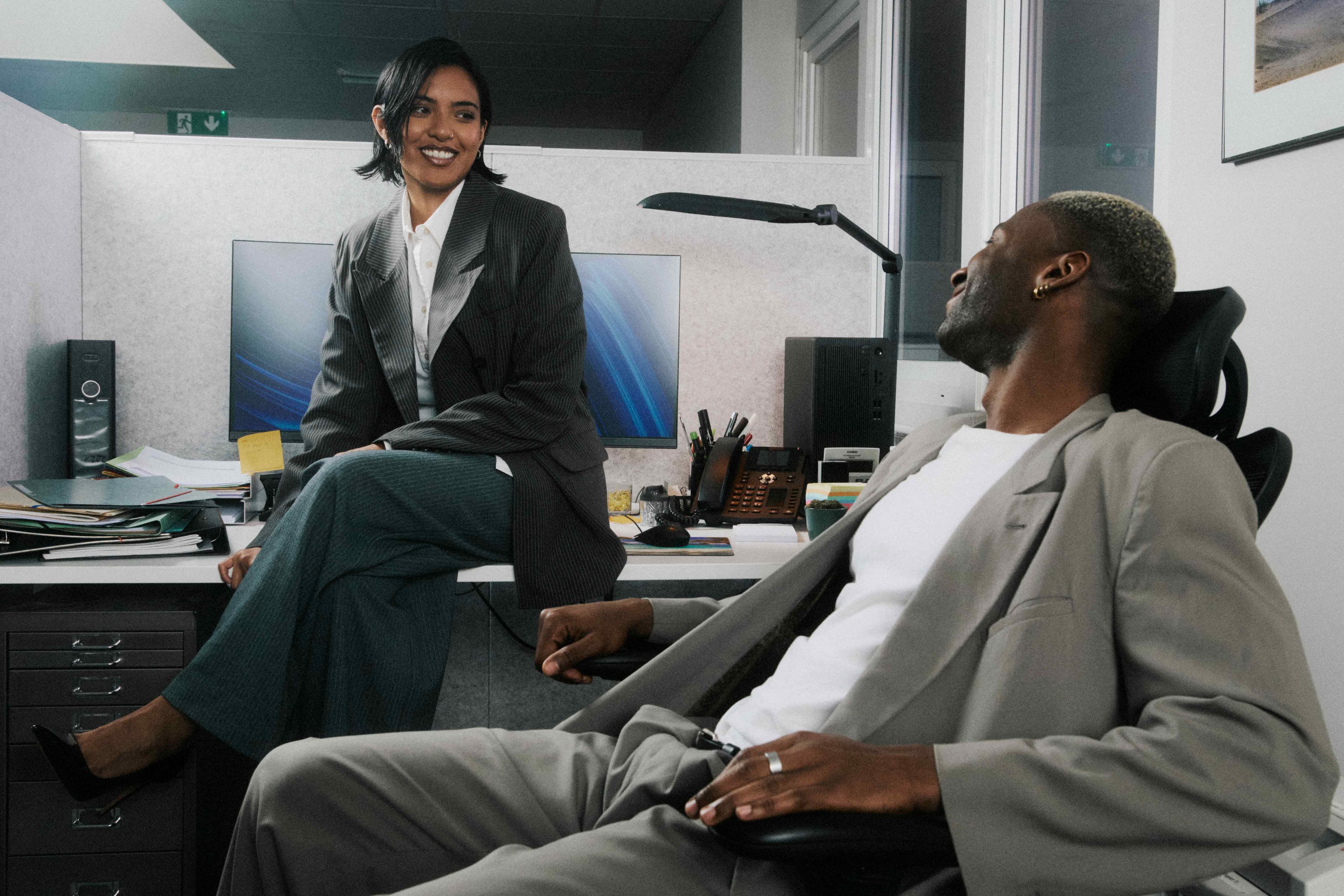 A woman on a desk edge laughing with a colleague.