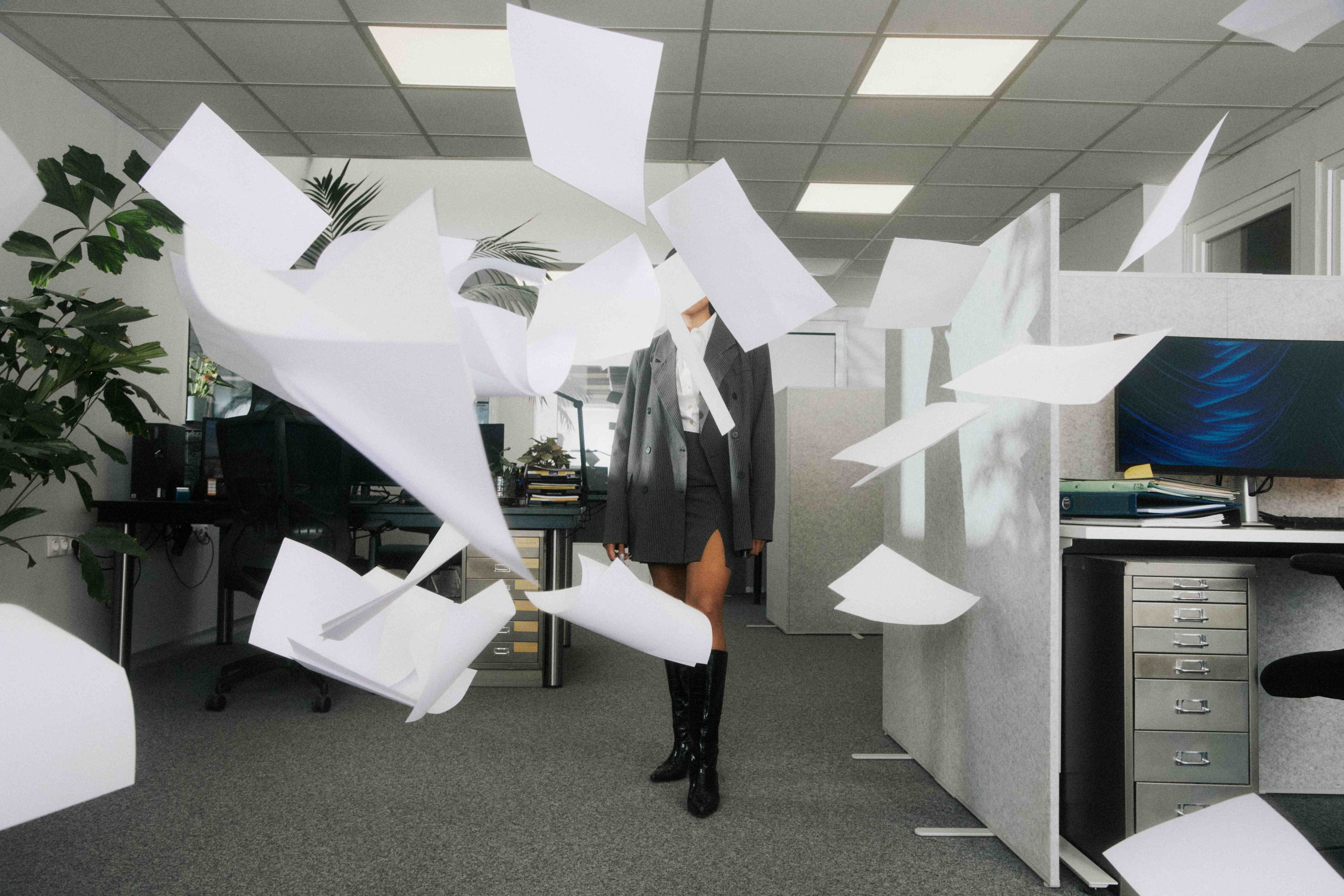 A composed person in oversized tailoring, unbothered while paperwork flies around the office